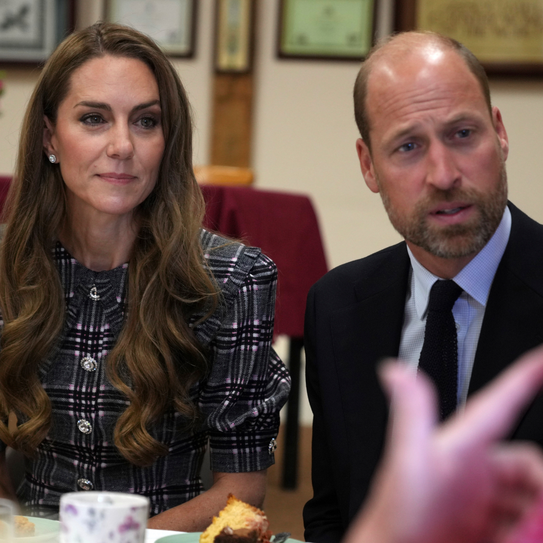 Kate Middleton wears a checked coat and has long wavy brunette hair while sitting next to Prince William