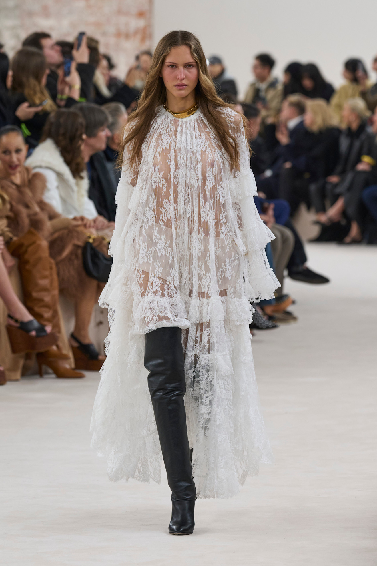 The Chloé’s A/W 2025 collection. The model wears a full lenght sheer white lace dress and black knee high boots. This image was taken from Launchmetrics.