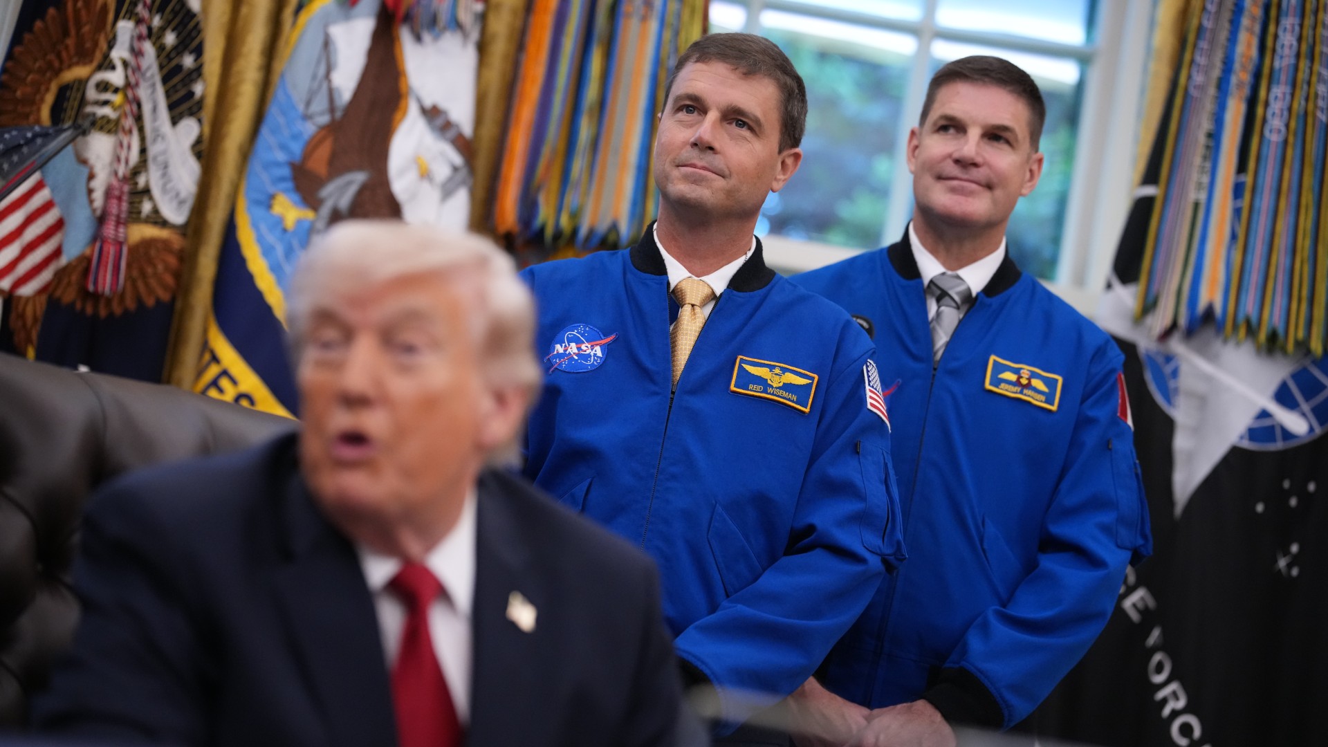 four people in blue flight jackets stand behind an old man at a desk in a grotesquely decorated office