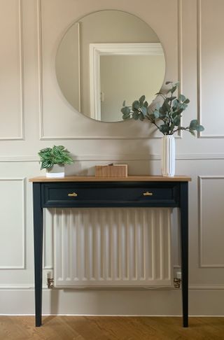 Image of a foyer with white walls that have wall molding on them. There is a small navy console table with two white vases on them with green plants. There is also a circular mirror hanging over the table