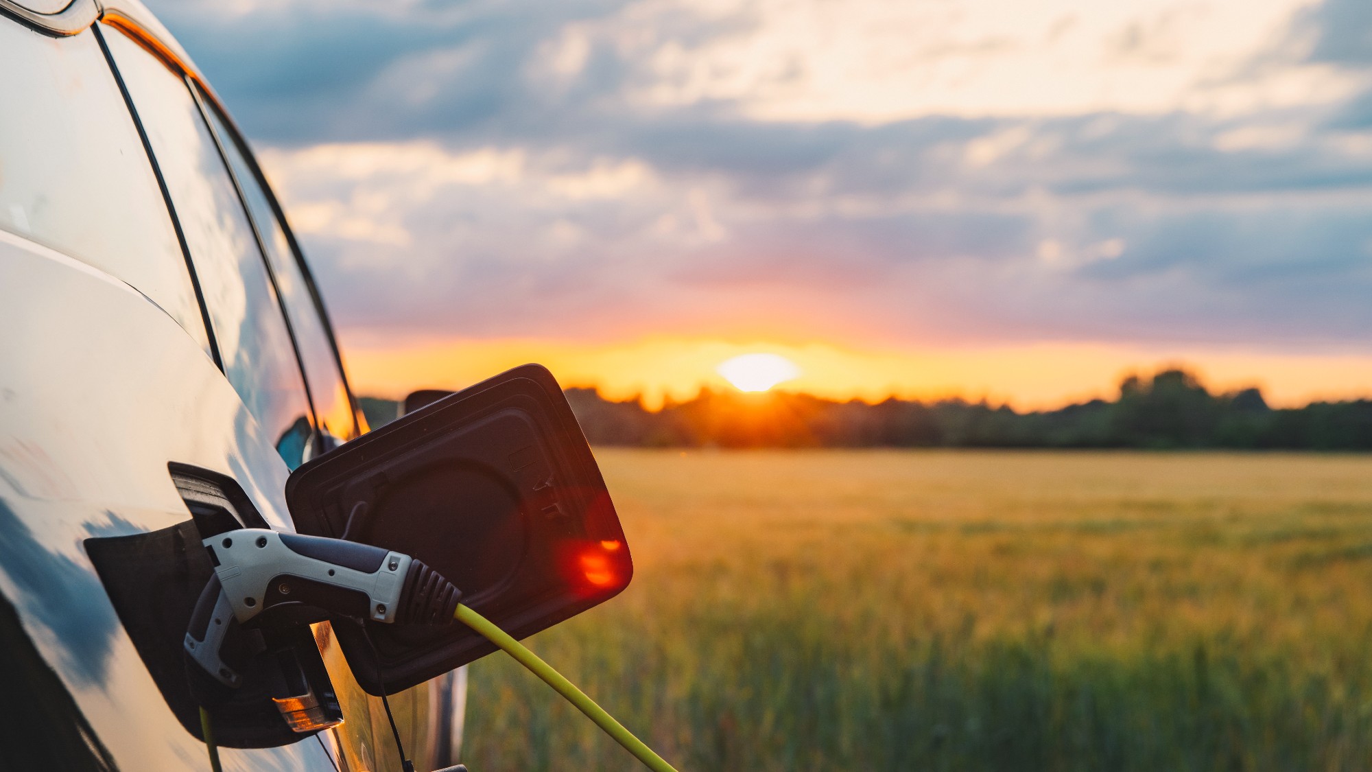 car charging port open with a charging cable attached. the car is in an open field and the sun is setting on the horizon