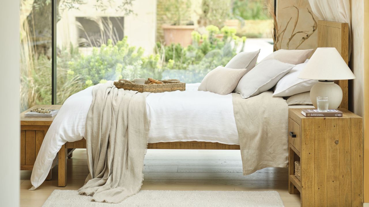 A wooden bed frame dressed in neutral coloured bedding in a room with a glass wall and a garden beyond