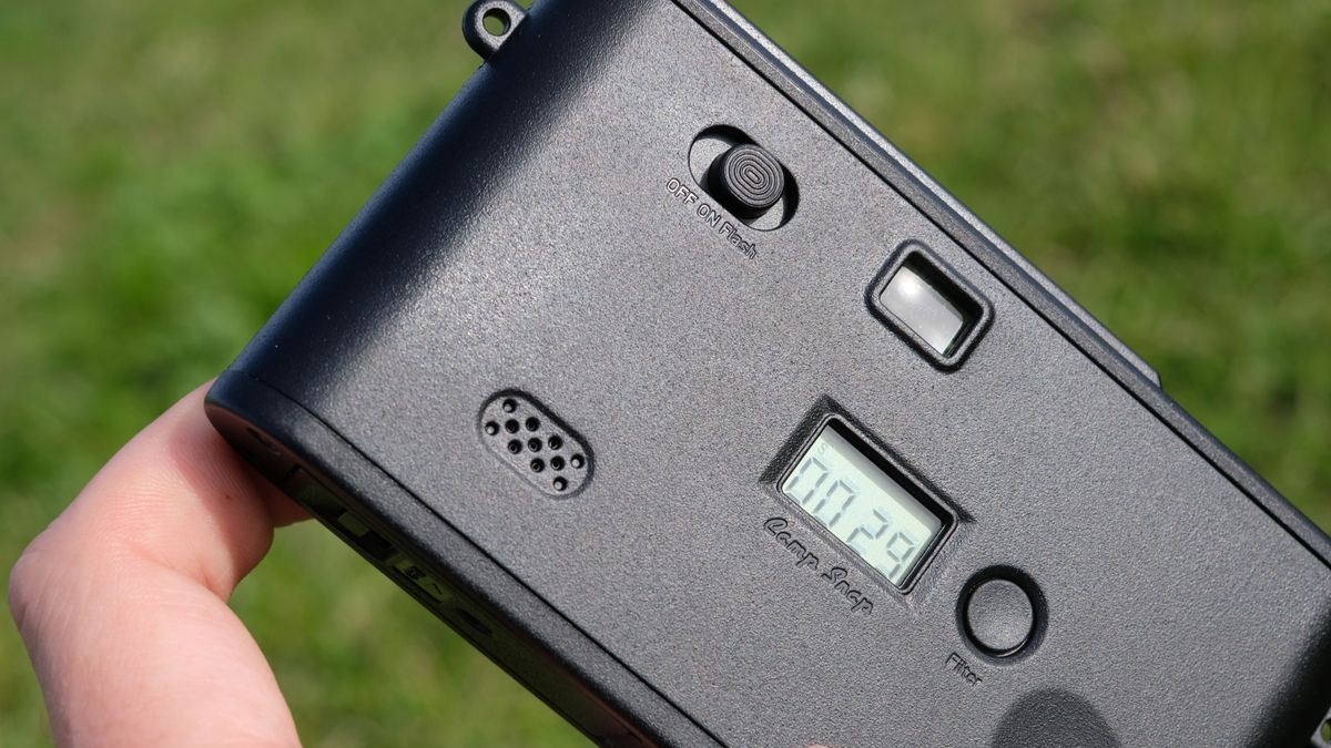 Photograph of man's hand holding Camp Snap 2 digital camera, in yellow colorway, rear view showing shot counter LCD, speaker, filter button and power switch