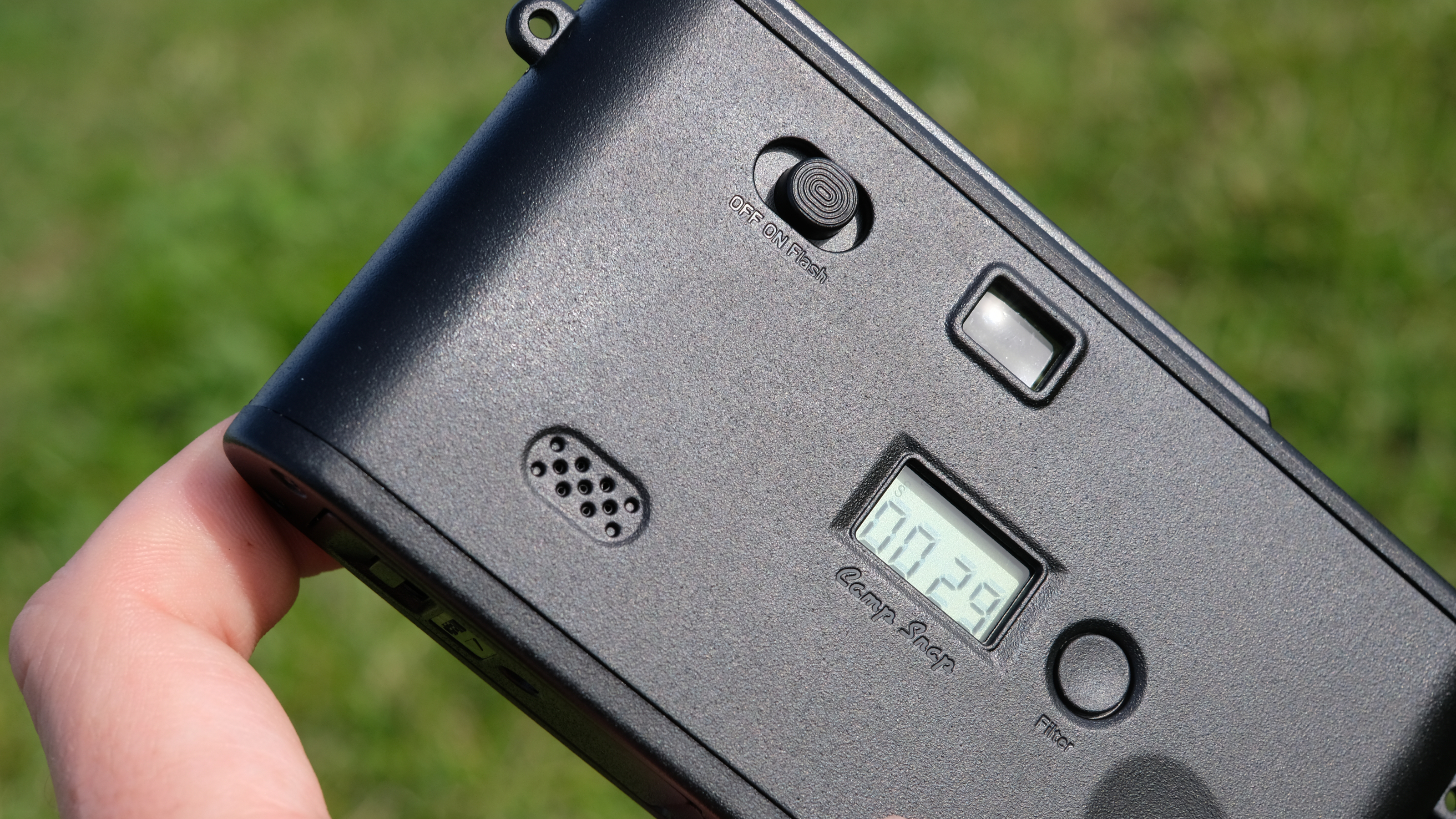 Photograph of man's hand holding Camp Snap 2 digital camera, in yellow colorway, rear view showing shot counter LCD, speaker, filter button and power switch