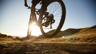 Is bike riding better for you than walking? Close up image of man's legs on bicycle, with mountains and sun flare in the background.