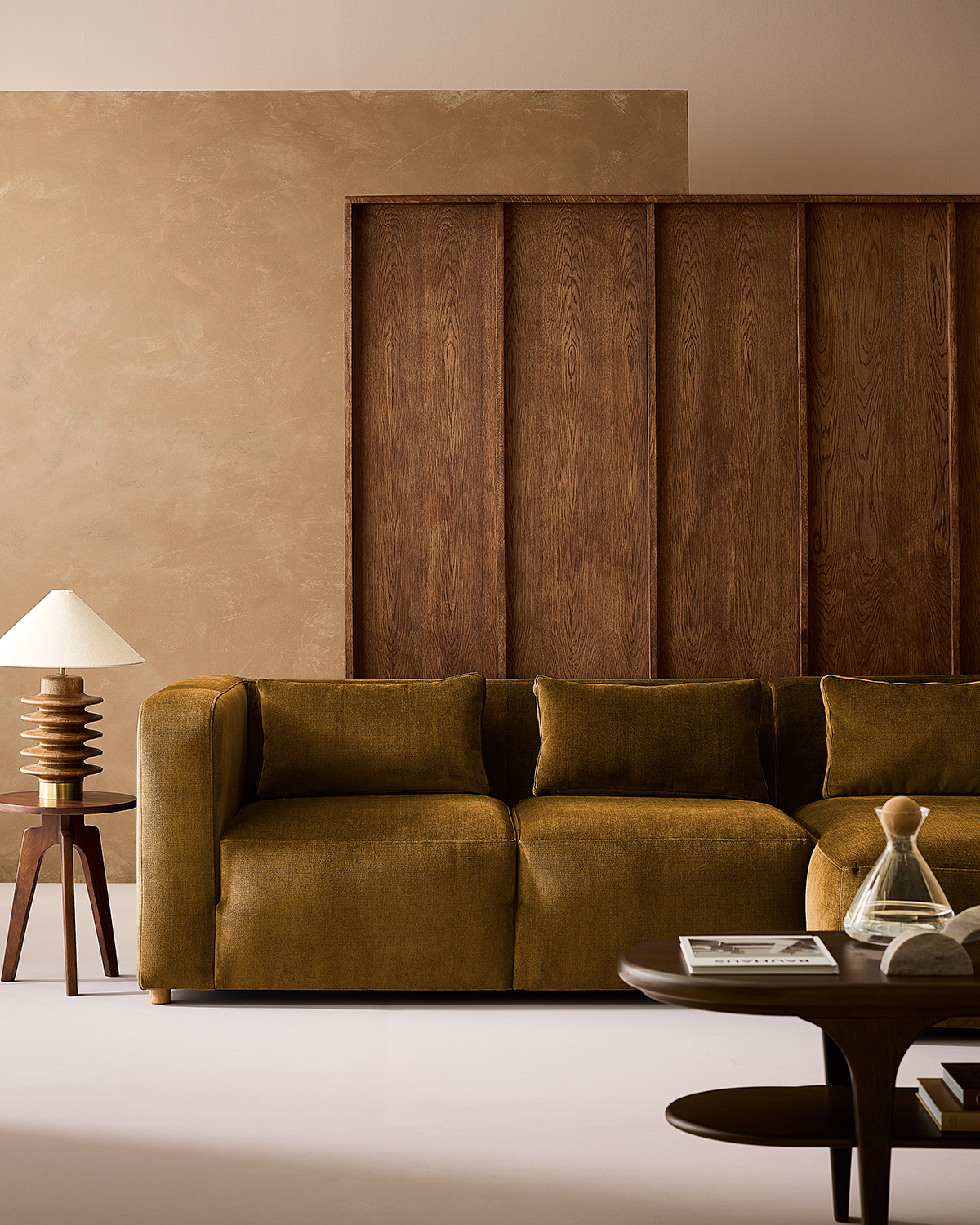 Image of a living room with beige walls and dark wooden paneling. There is an olive green, velvet sofa and a small wooden side table with a wooden accent lamp on it.
