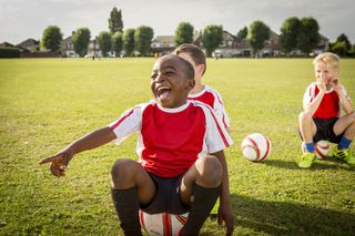 Boy in football kit sitting on a football on a pitch, smiling and laughing, there's a boy in the bakground also perching on a football