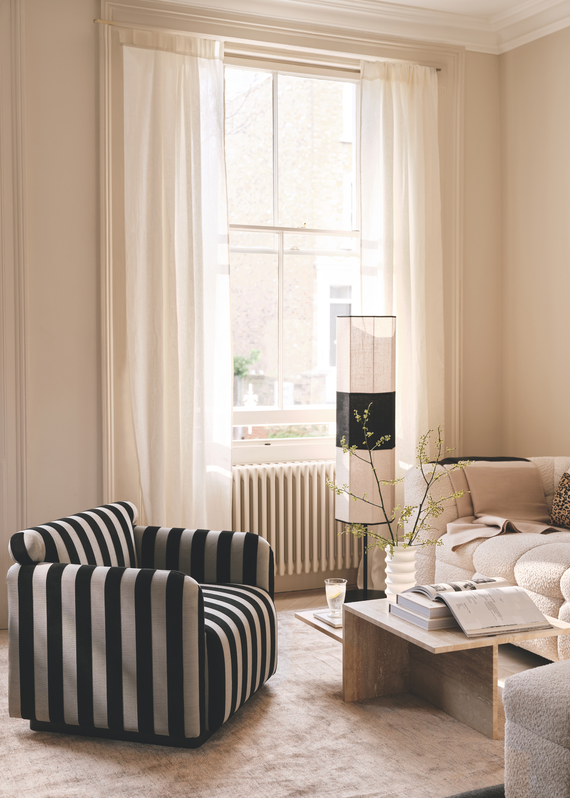 A bright living room with a striped accent armchair, a boucle couch, a marble side table with a vase, a glass and some books beside a standing black and white lamp