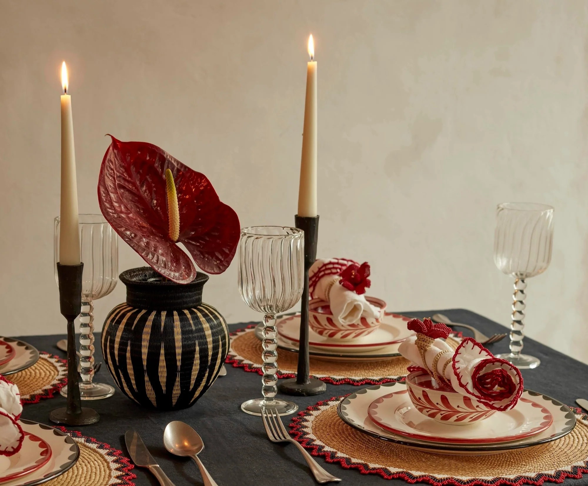 A beautiful dining table decorated with rattan placemats, red and white napkins, organic-shaped glasses, and lit tall cream candles, along with a red flower.