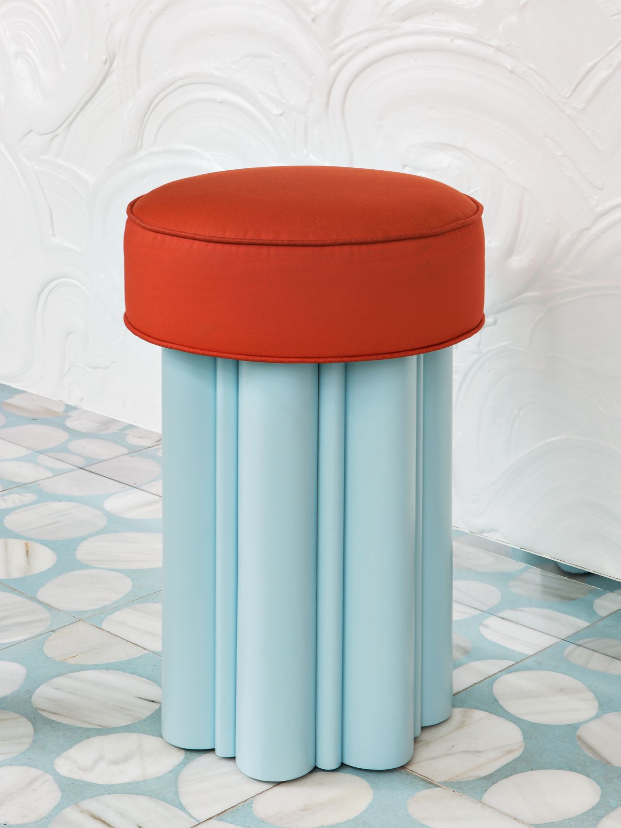 An upholstered stool with red seating and light blue base sits on top of abstractly decorated tiles in light blue and white.
