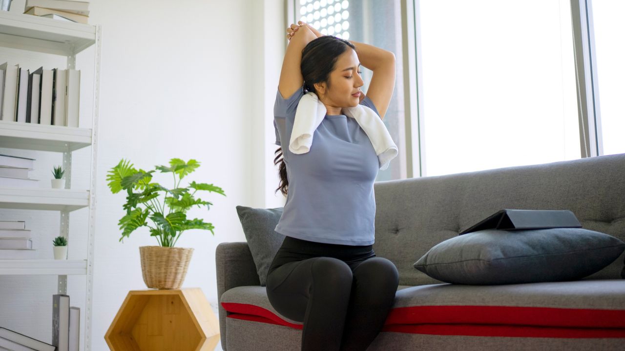 Woman in a living room sitting on her couch with a towel around her neck doing a triceps stretch