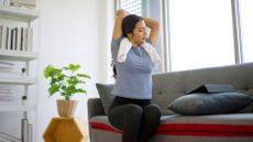 woman in a living room setting sat on her sofa with a towel around her neck doing a triceps stretch.