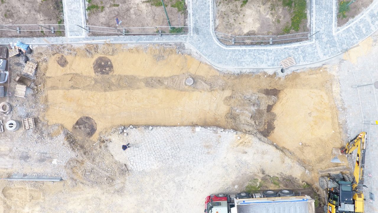 An image of the burial site taken from a drone. Here we see an aerial view of a street dug up and cordoned off with some metal fences. There is a digger in the bottom right corner.
