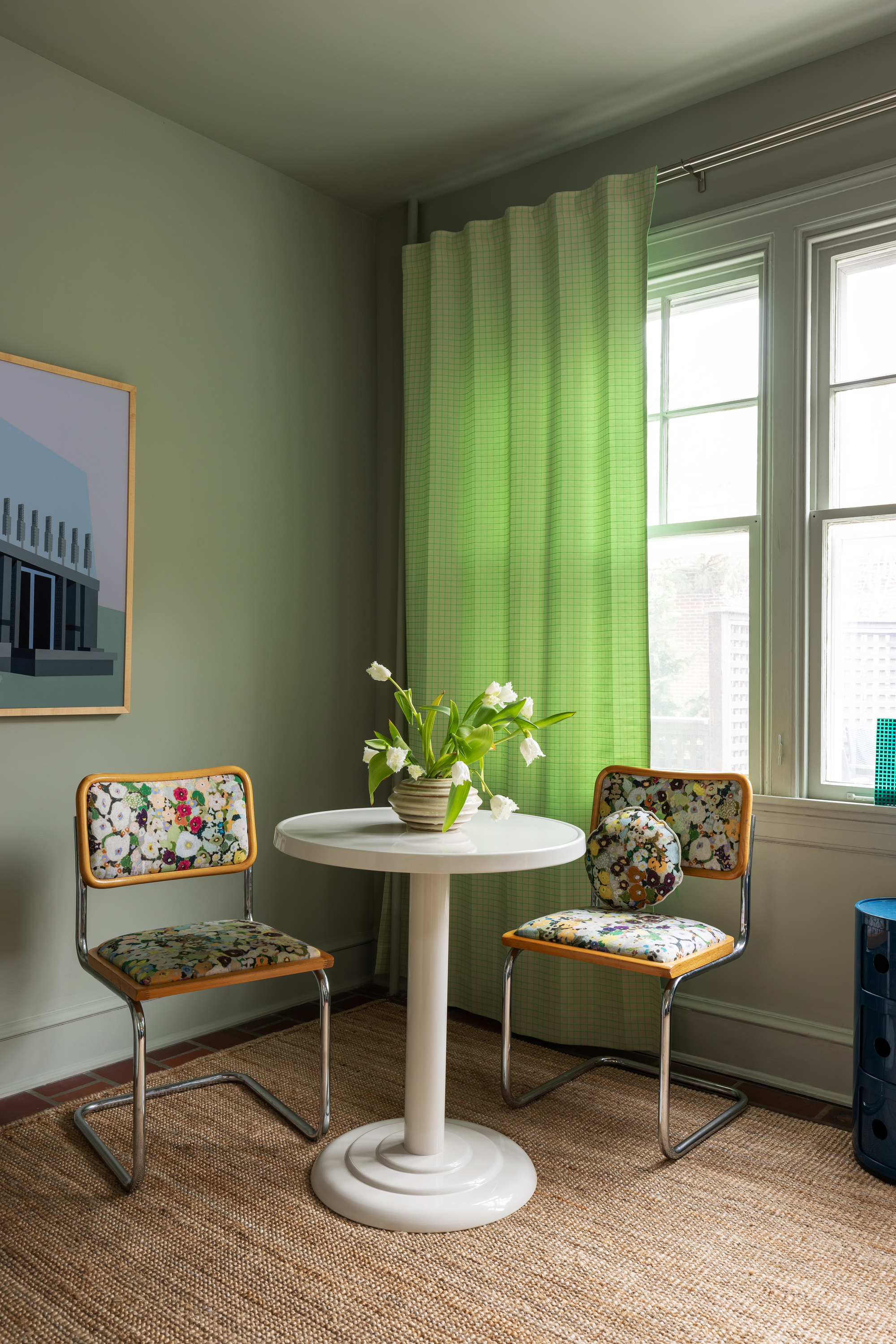 Image of a dining corner in a green room with a white table and two floral, retro chairs. There is a window with a bright green curtain.
