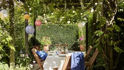 Pergola ideas are so pretty. Here is a wooden pergola with vine leaves around it, festoon lights and colorful lanterns above it, a white tablecloth with plates on it and wooden chairs around it