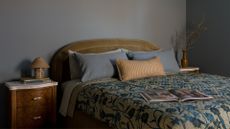 A beautiful blue bedroom with a wood side table a small table lamp. blue pillows, a striped bolster, a patterned quilt, and a curved green headboard