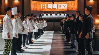 Two rows of chefs (the White Spoons, left, and the Black Spoons, right) stand facing each other, with a circular dining table in the back and a screen reading "1:1" with a Korean phrase, in 'Culinary Class Wars.'