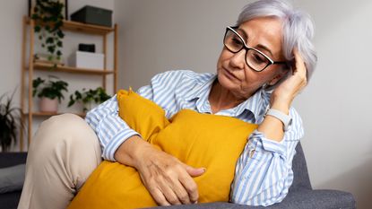 An older woman looks serious as she sits on her sofa, holding a pillow to her chest.