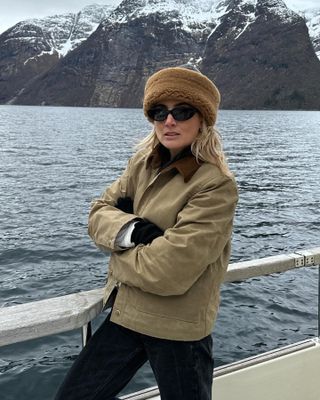 British style influencer Lucy Williams poses in front of a snowy mountain in Norway wearing a Toteme shearling beanie hat, black oval sunglasses, and a tan barn coat