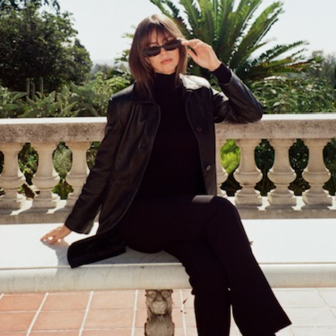 Kathryn Zahorak sitting on bench wearing sunglasses, black turtleneck, leather jacket, and black pants