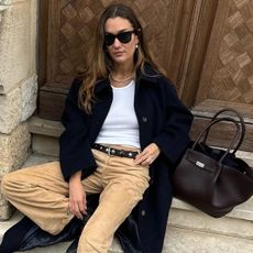 Woman wears white shirt, navy coat, tan couduroy pants, black belt, and leather bag while sitting down on Parisian street steps.