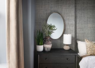 A close up of the image of the bedside area with bedside drawers, a mirror, and grasscloth wallpaper behind