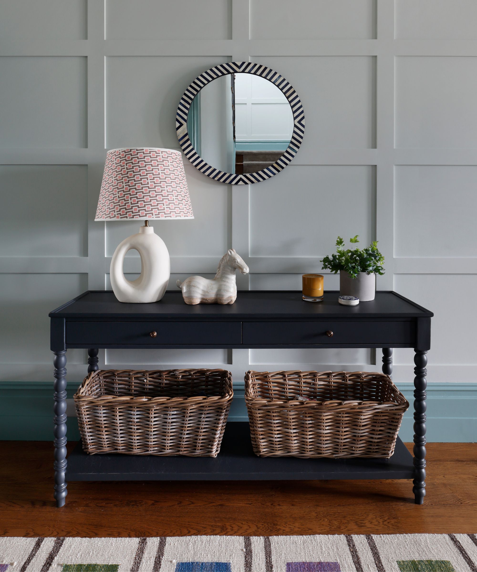 Entryway with wooden panelling on the wall painted in a pale blue, a dark console with bobbin legs, a round mirror, storage baskets underneath, a round white lamp, and a plant on top.