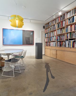 A human shadow in on the floor in a room next to a wall shelf of books on the right. On the left is a white table with mathcing chairs.