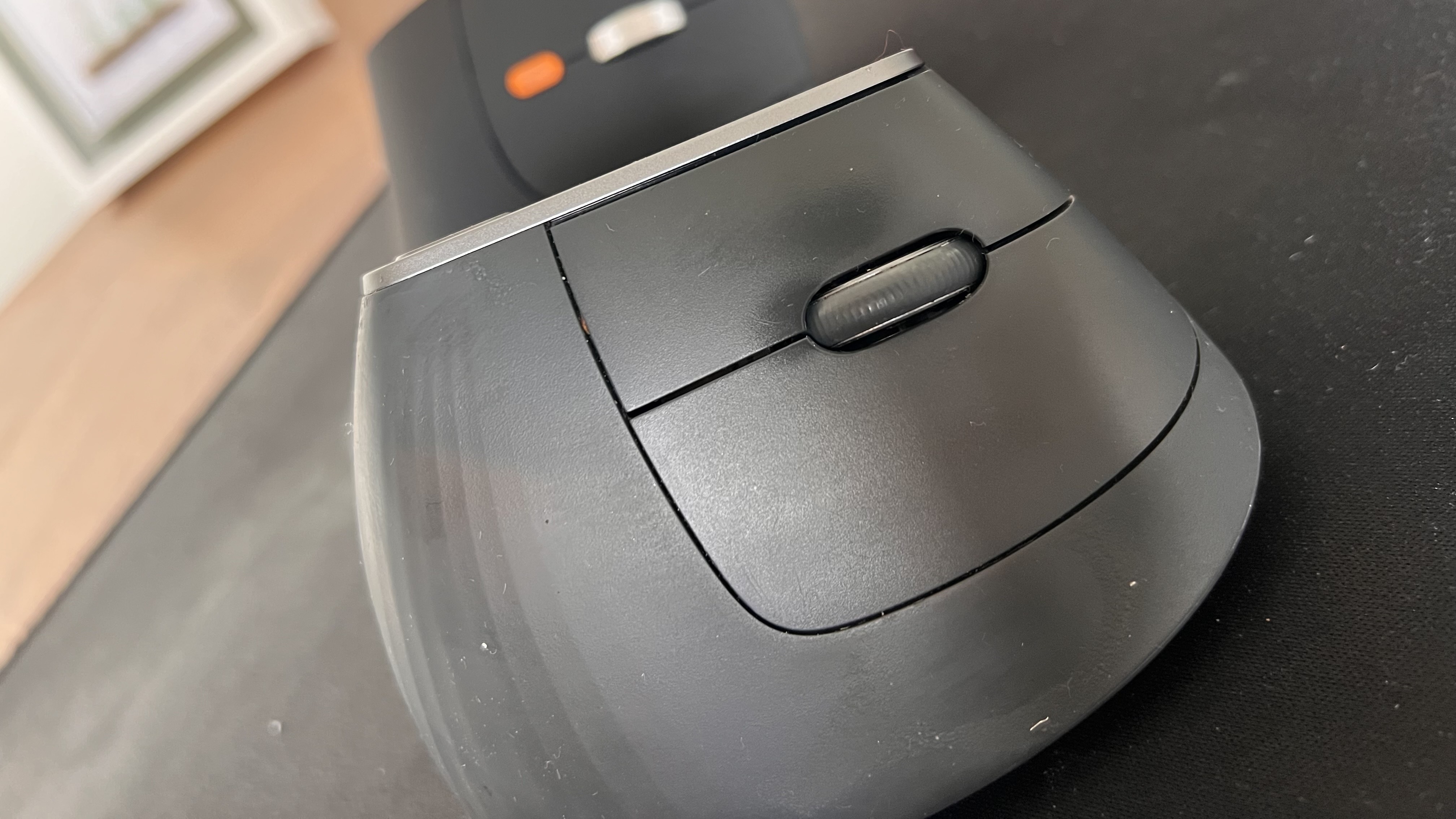 Close-up front view of the Logitech MX Vertical mouse showing the top scroll wheel and right-click buttons, with the ProtoArc EM11 Pro partially visible blurred in the background.