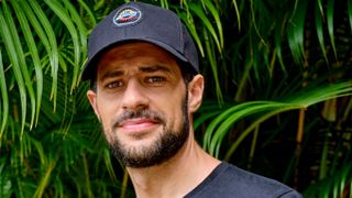 Dale (David Avery) stands in front of some green palm fronts, wearing a navy blue baseball cap with the Island Warrior logo on it and a dark blue t-shirt. He has a troubled expression on his face.