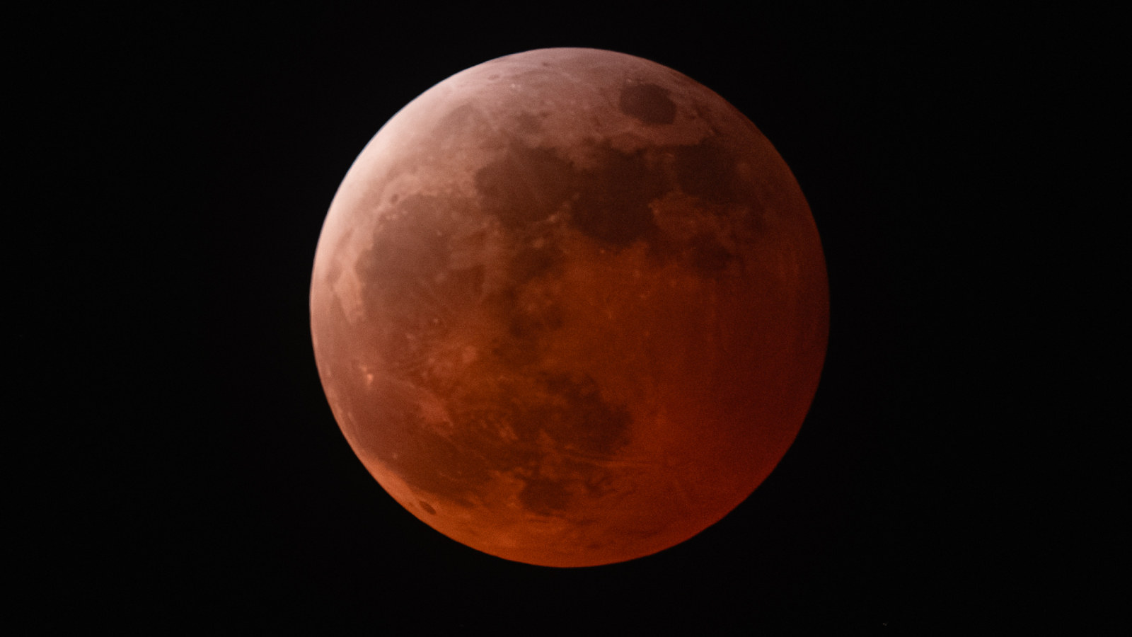 An orange-red full moon is pictured against a black night sky. The upper left of the lunar disk is relatively bright. Vast basaltic plains known as lunar maria can be seen dominating the left half of the moon's surface.