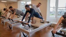 Instructor James Shaw teaches at a busy Pilates class. We see four clients on reformer machines in a plank position. Shaw is helping position one of the client's head and calves properly, standing next to her to perform adjustments. 