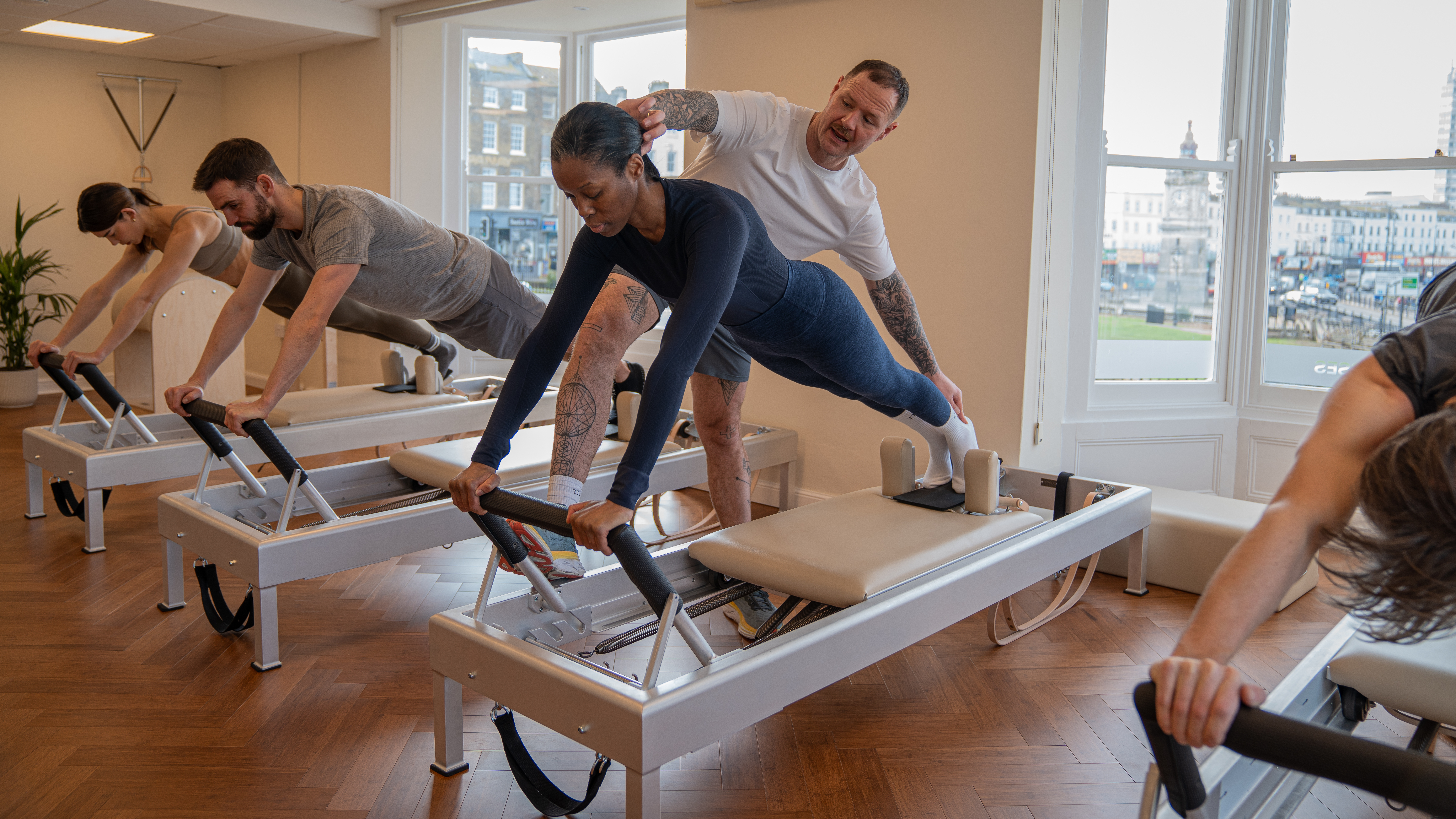 Instructor James Shaw teaches at a busy Pilates class. We see four clients on reformer machines in a plank position. Shaw is helping position one of the client's head and calves properly, standing next to her to perform adjustments. 
