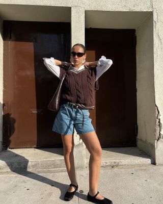 Aisha Farida wears denim shorts, a sweater-vest, Mary Janes, and a white button-down shirt.