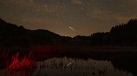 A meteor is pictured streaking through a starry sky above a hilly tree-lined horizon. A lake is visible at the bottom of the image, reflecting the sky above.