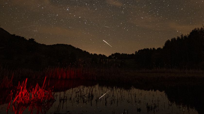 A meteor is pictured streaking through a starry sky above a hilly tree-lined horizon. A lake is visible at the bottom of the image, reflecting the sky above.