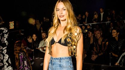 Margot Robbie attends the Chanel 2024 Cruise show in LA wearing wide-leg jeans, and a gold vest. 
