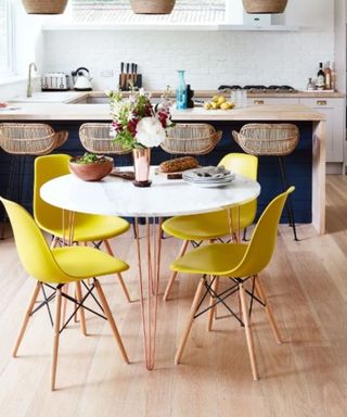 A kitchen with a white dining table with yellow seats, and a dark blue kitchen island behind it with rattan seats, plus white walls and wooden floors