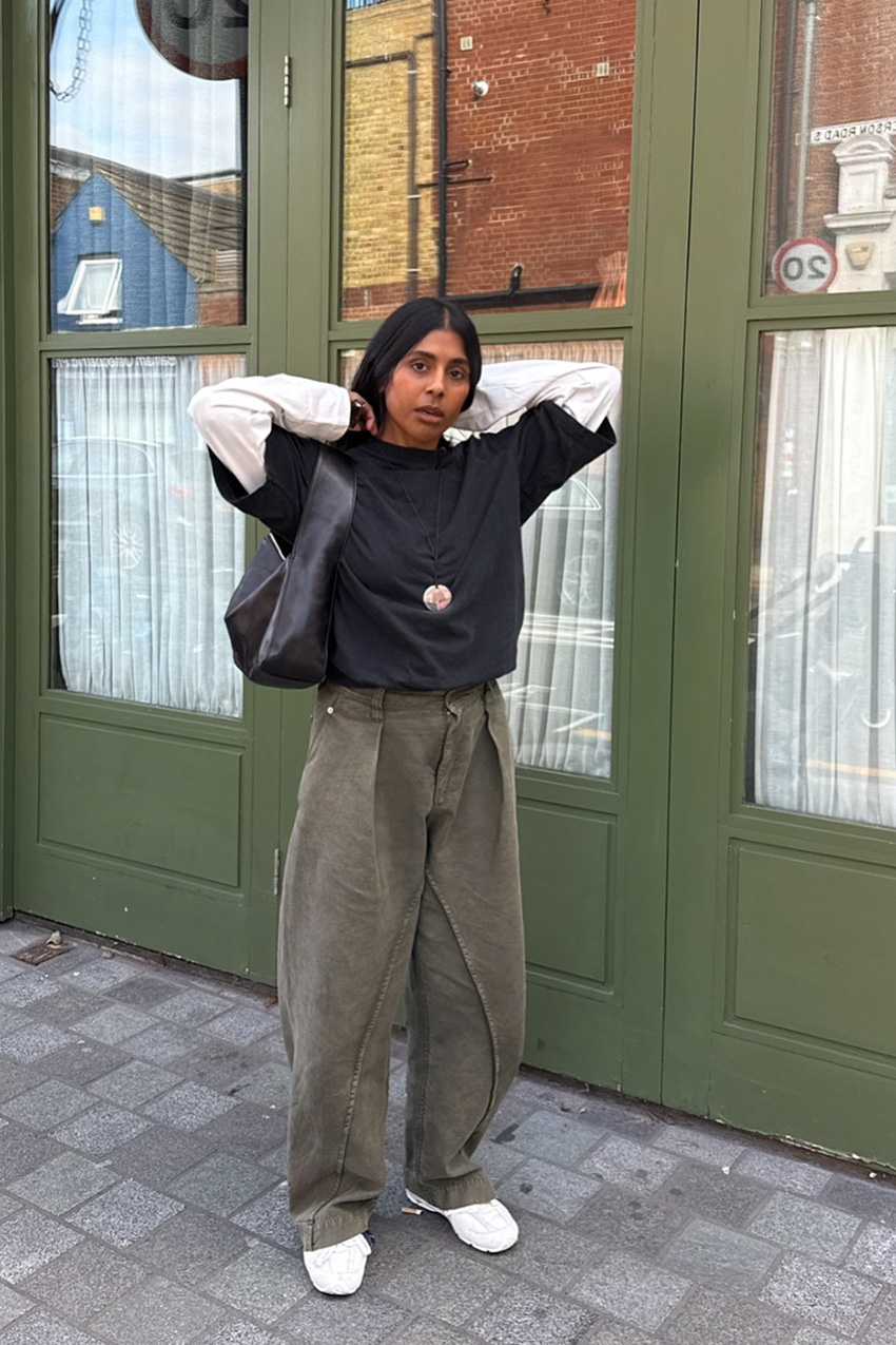Layering staples for spring 2026 is shown in a street style photo with a woman standing outside, while wearing a white t-shirt layered under an oversized black t-shirt styled with baggy green cargo pants, white sneakers, a black shoulder bag, and a cord necklace
