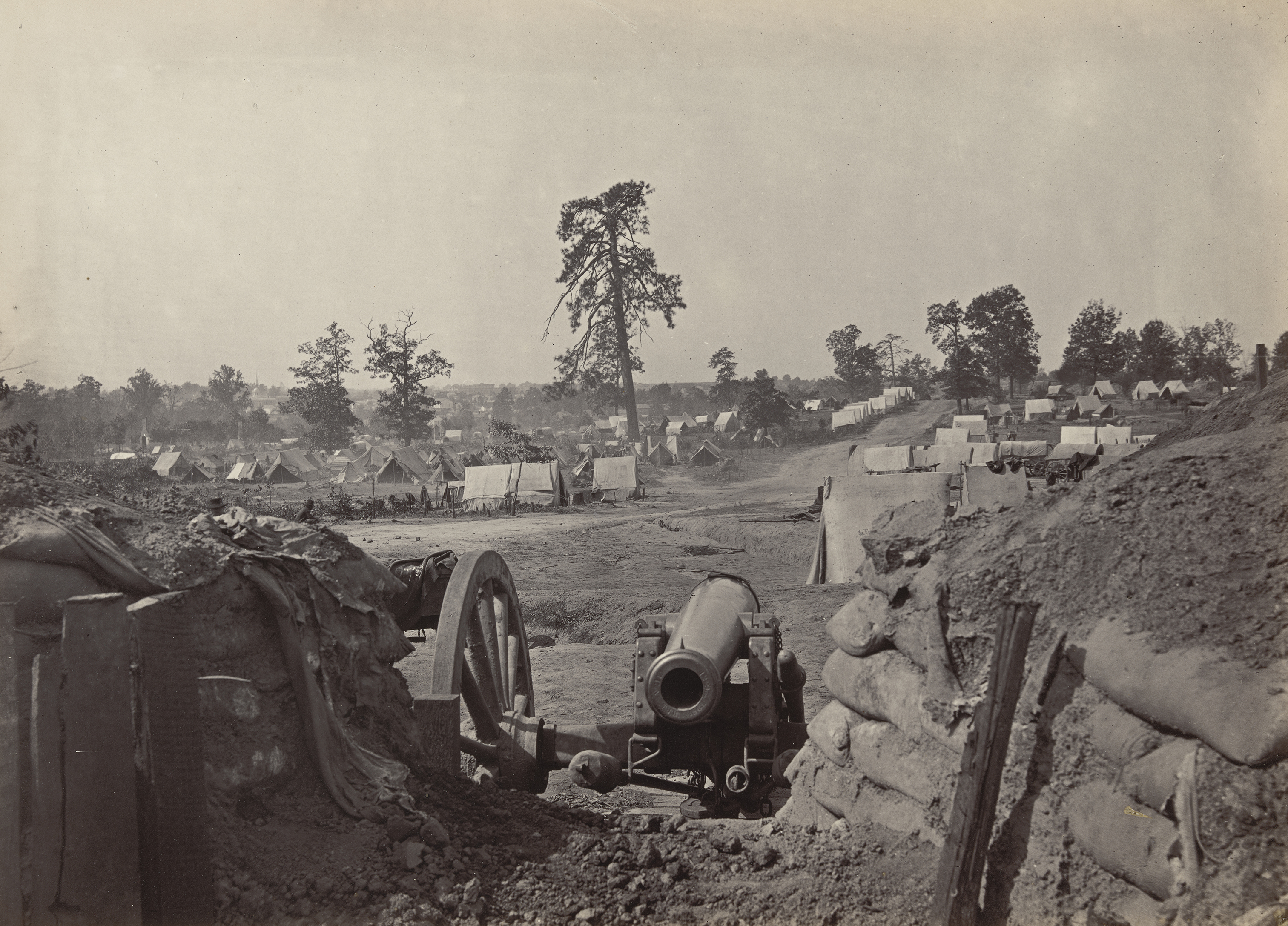 A heavy cannon sits positioned behind a low earthwork of sandbags, looking out over a sprawling military encampment filled with white tents.