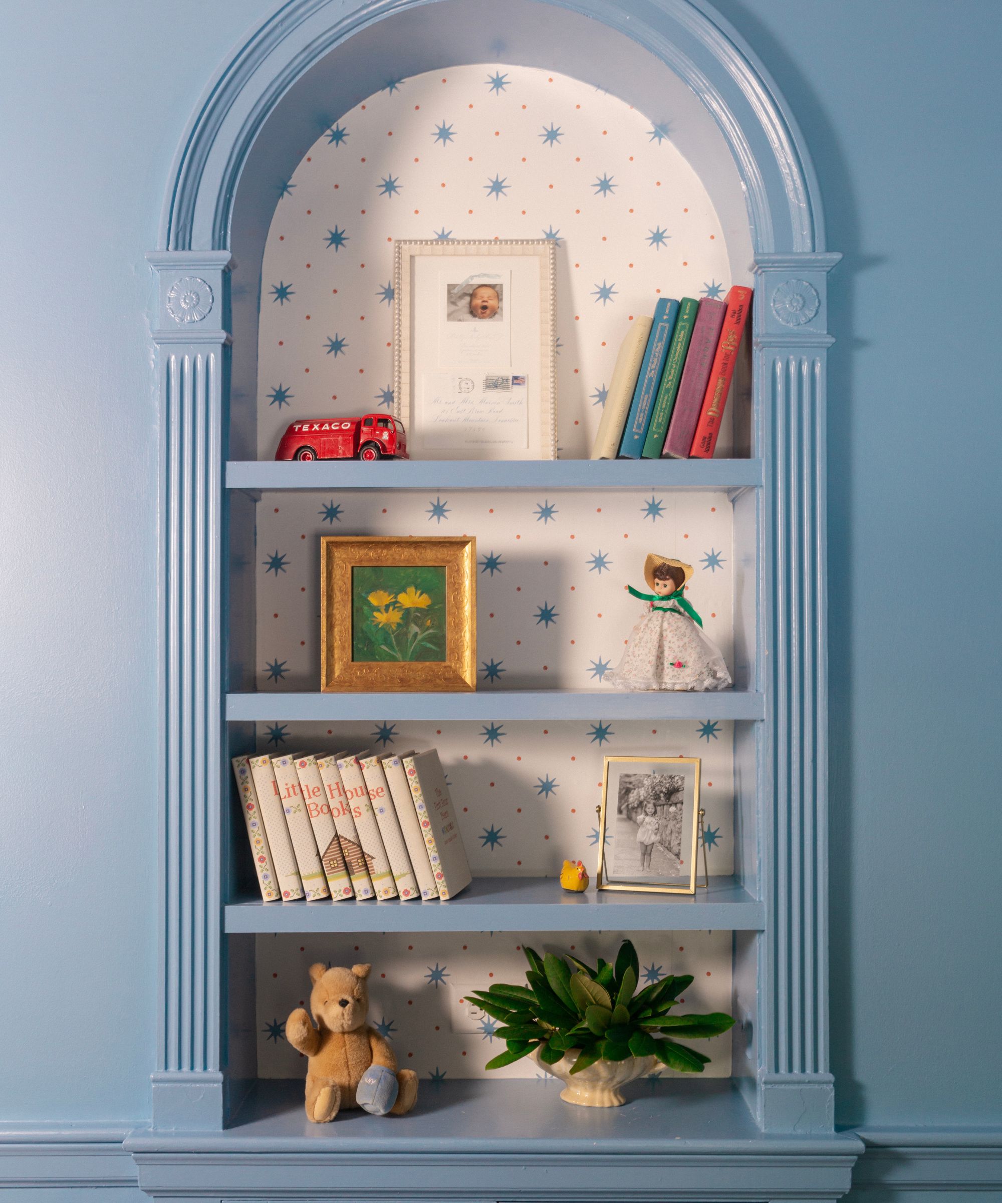 Blue painted alcove with a wallpapered background and vintage books and pictures along the shelves