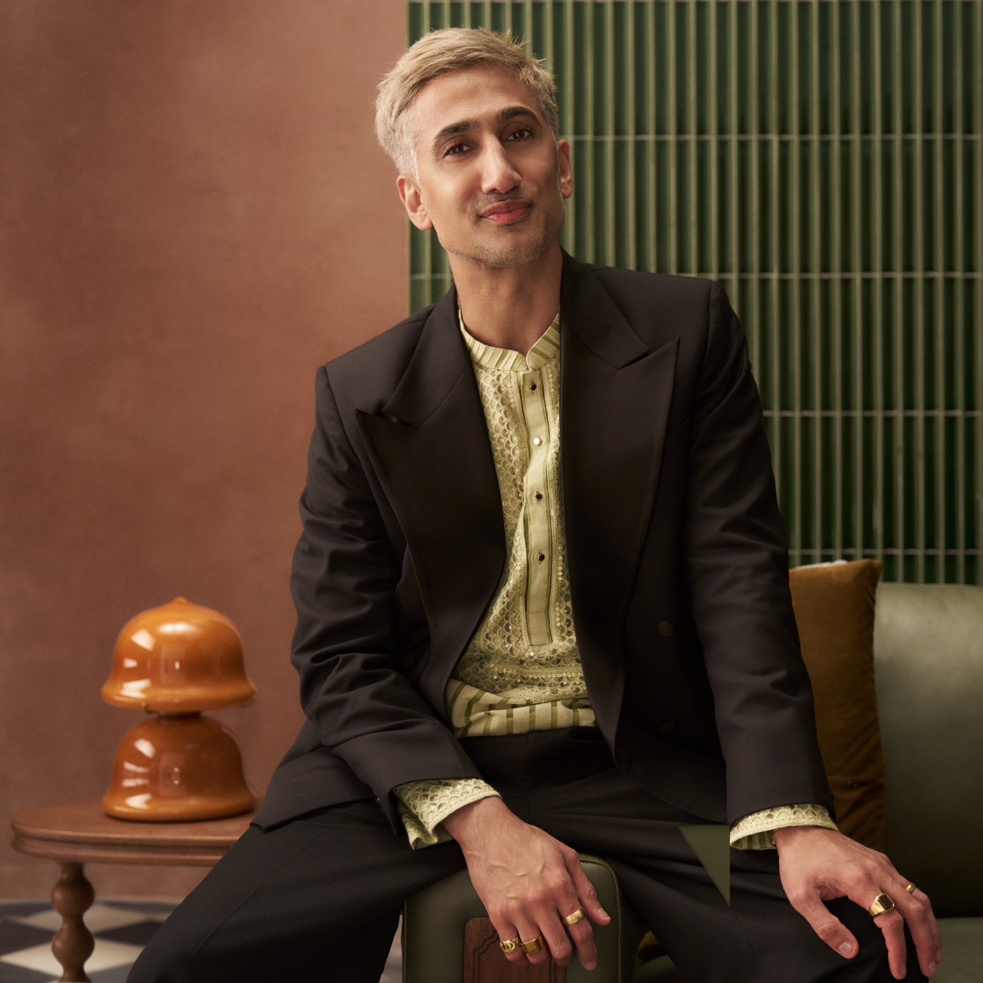 Tan France sittin on the armrest of a green leather sofa from his collection with Castlery with an orange glass table lamp on a side table behind