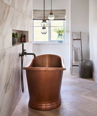 Copper bath, marble tiles on wall, wooden flooring, wooden ladder with towels, trio of pendants