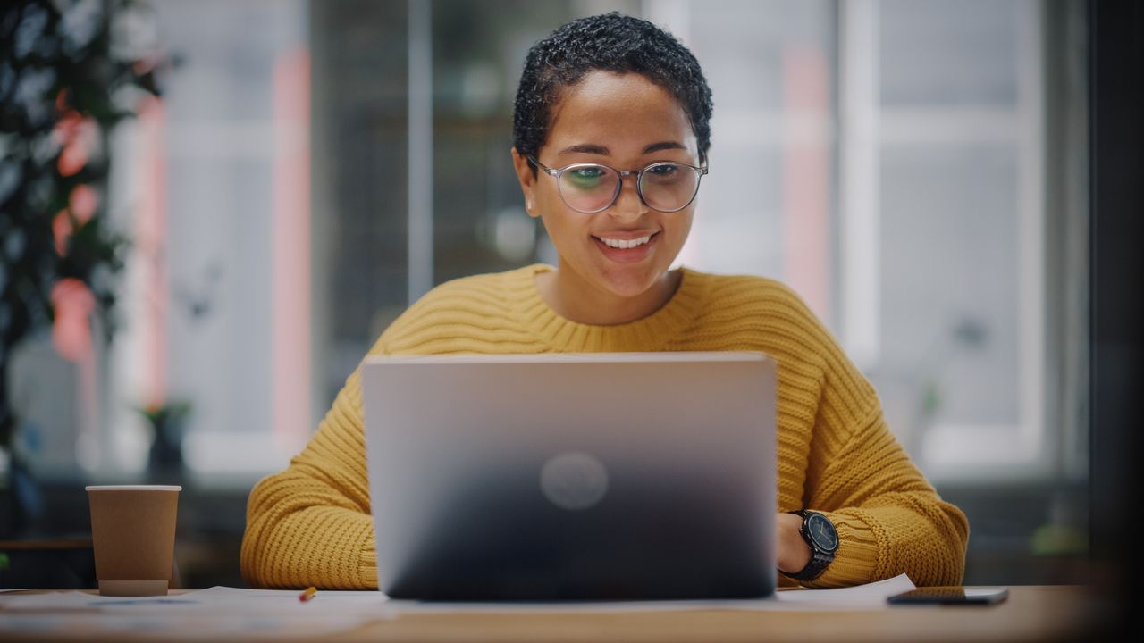 A woman smiles as she looks at her laptop at her dining room table.