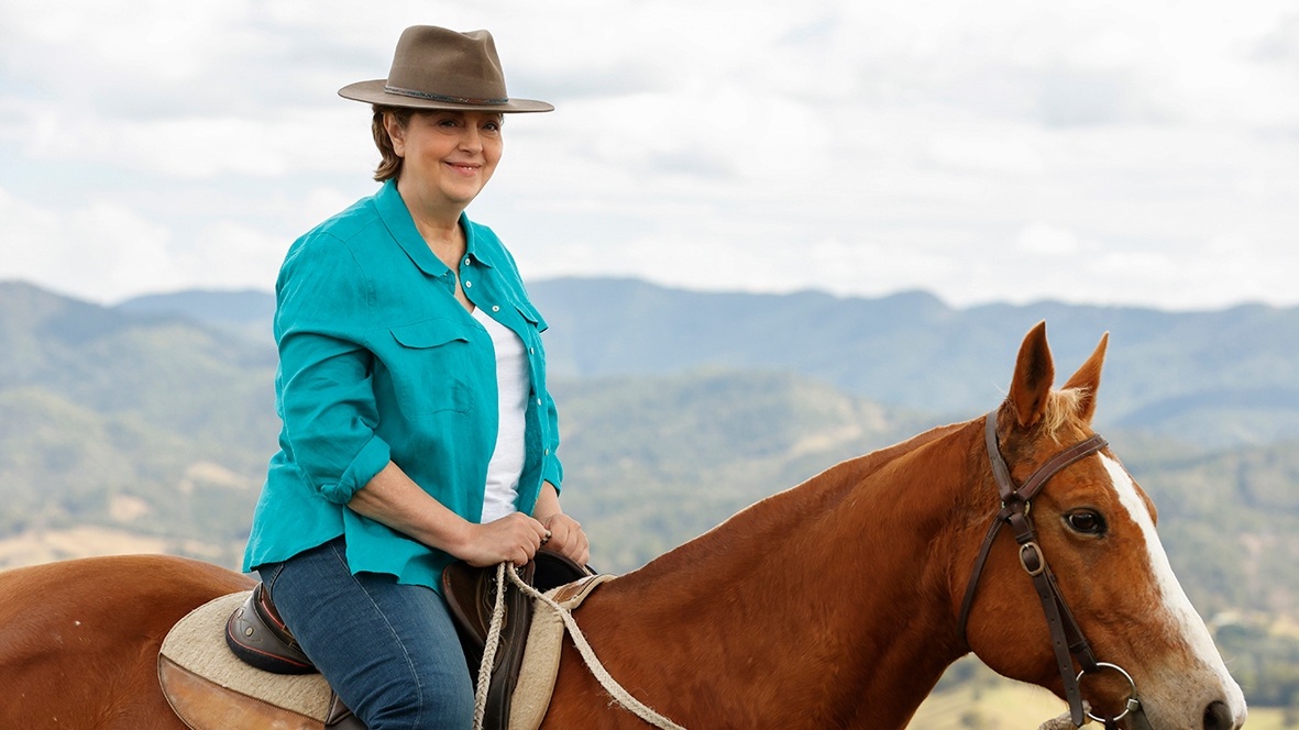 Joan on the back of a brown horse while weaing a turquoiuse shirt