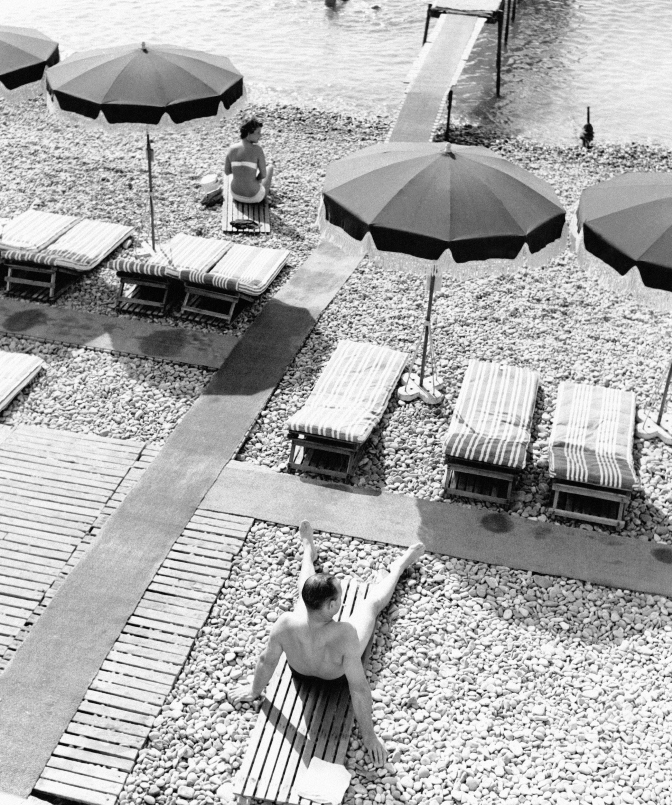 Two people sitting on a stony beach with an assortment of sun loungers and umbrellas