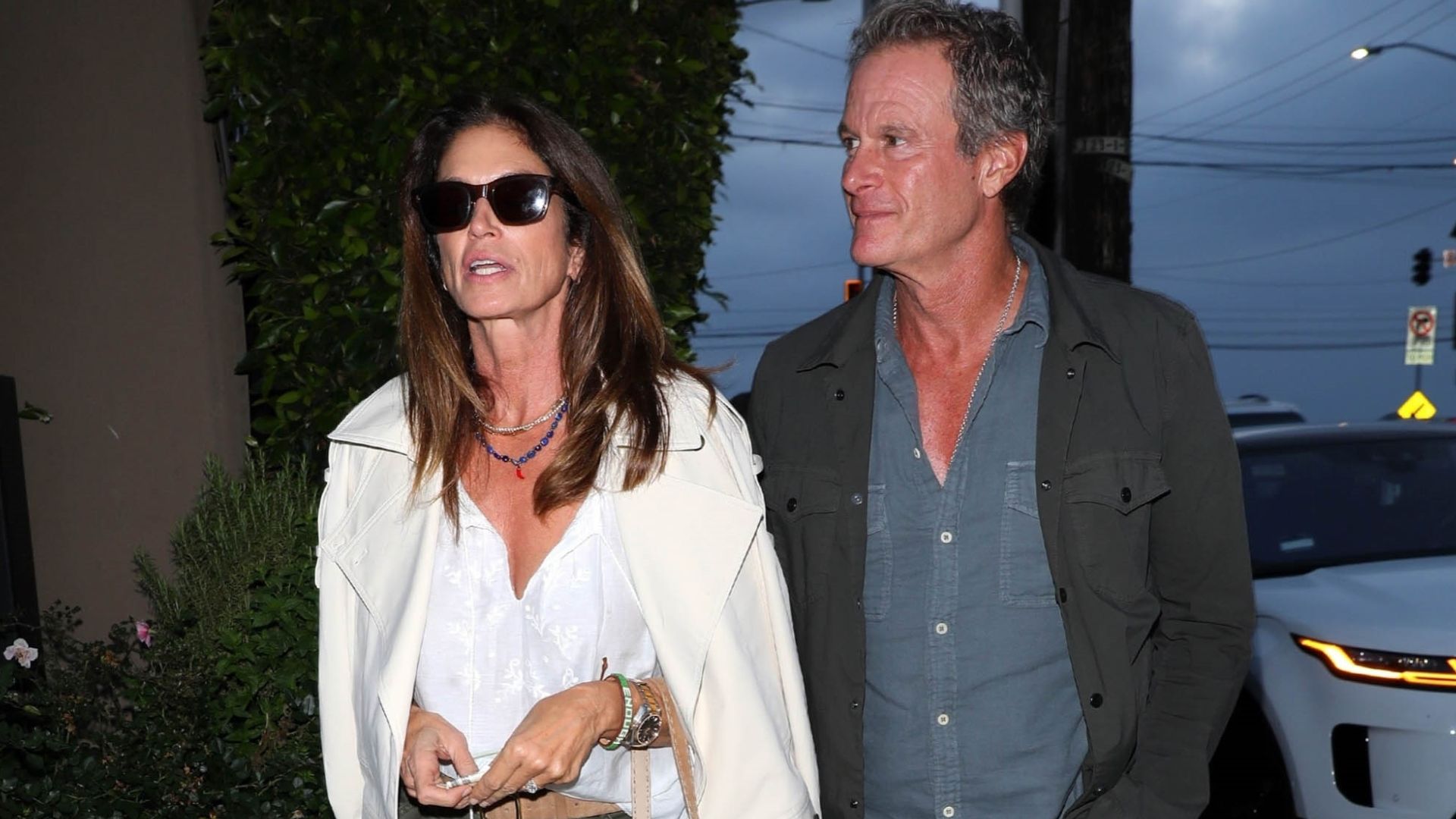 Cindy Crawford walks down the street wearing khaki jeans with beige suede sandals and a white blouse.
