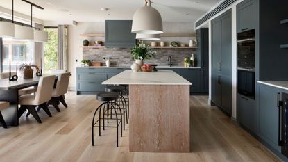 A dark blue fitted kitchen diner, with a large wooden island. Industrial style black barstools pulled up to the island, with a large black dining table behind it, with white upholstered chairs. 
