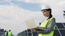 A woman in a high-vis vest and hard hat holding a laptop outside in a solar farm.
