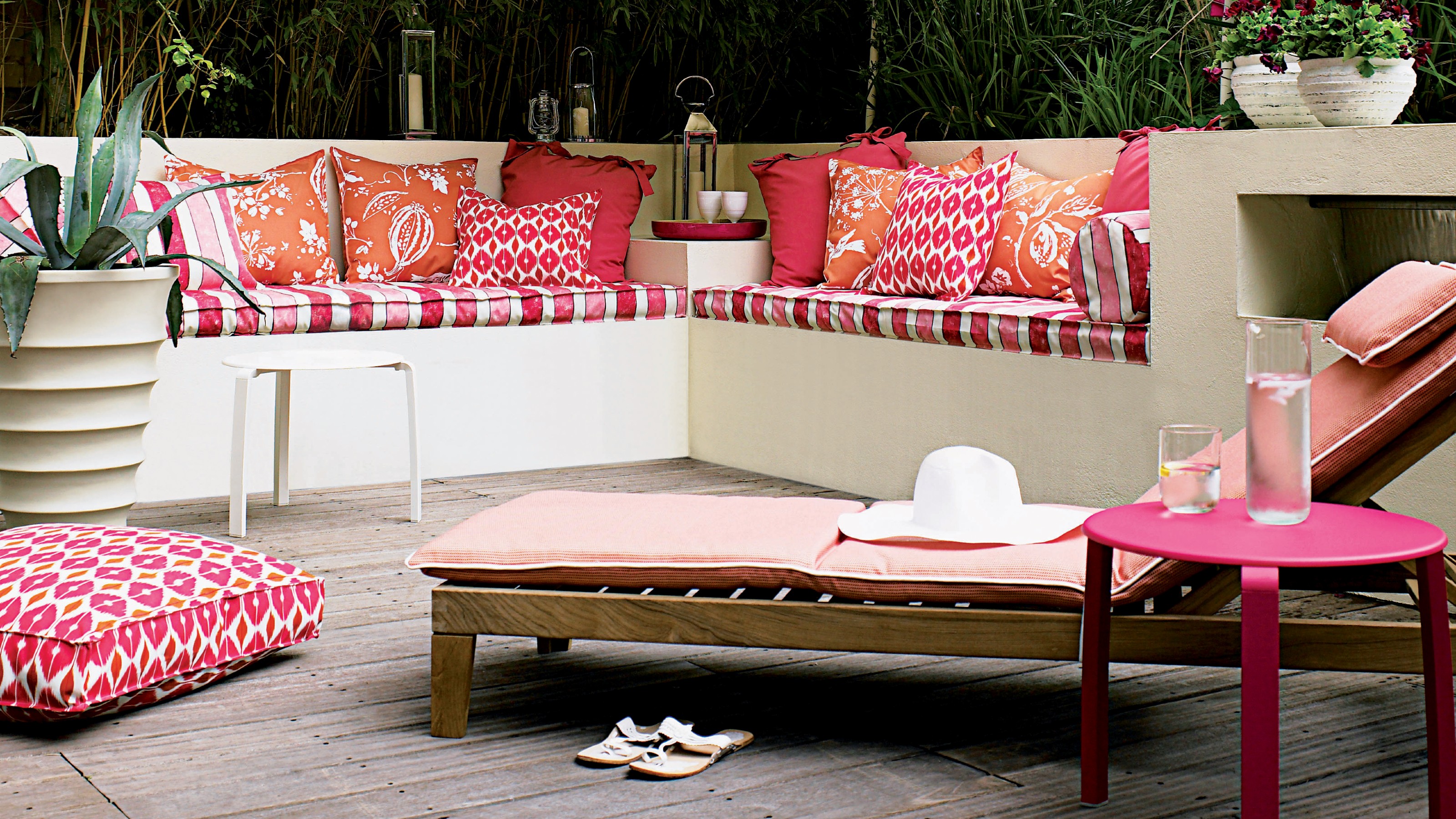 A garden with a wooden adjustable recliner sun lounger with a pik cushion pad and built-in bench seating adorned with patterned pink cushions